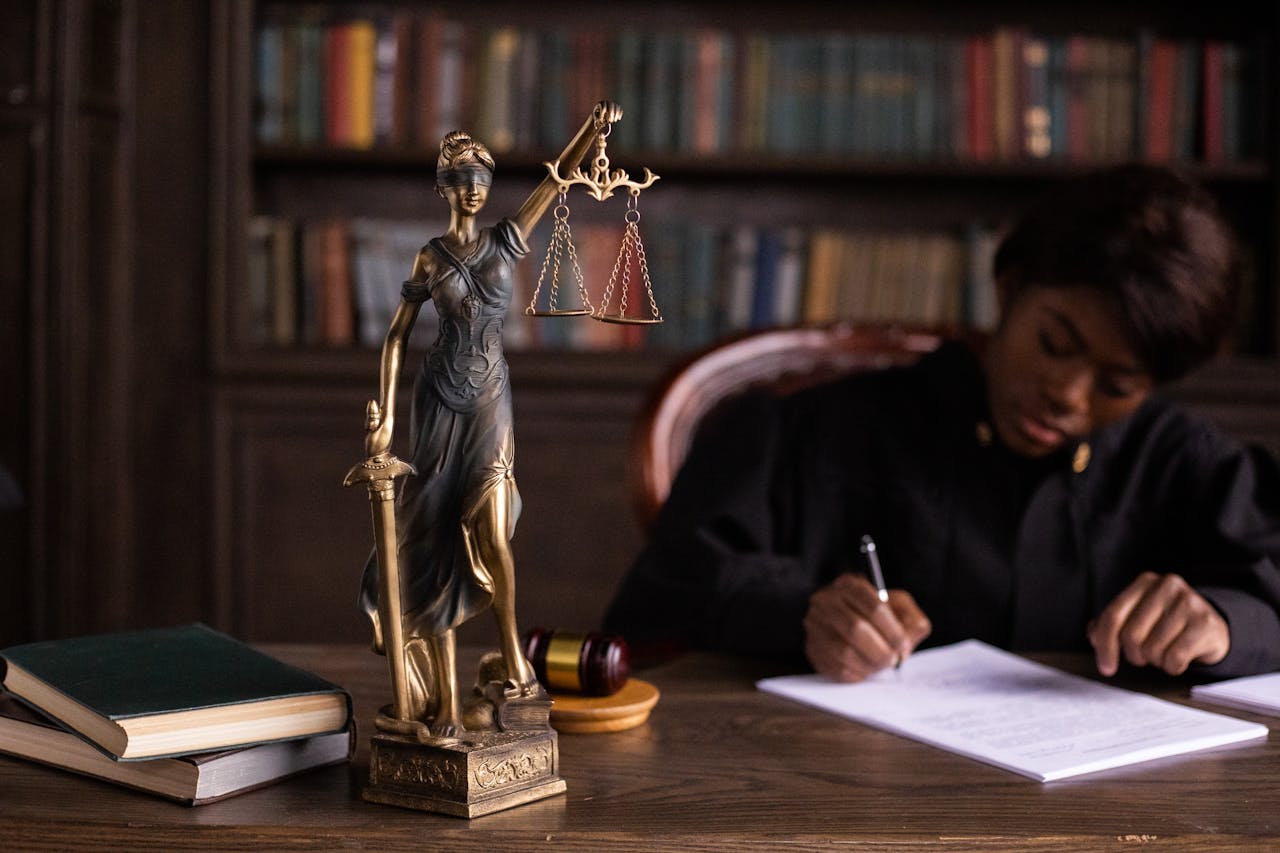 who-we-are A focused judge writing on documents beside a Lady Justice statue in an office.